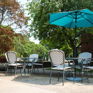 A serene outdoor patio area with tables and chairs under a teal umbrella, surrounded by lush greenery.