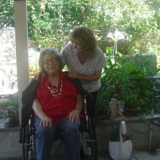 An elderly person in a wheelchair is being assisted by a caregiver in an outdoor garden setting.