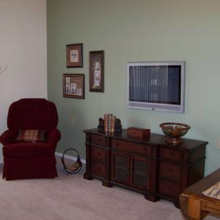 A cozy living room featuring a maroon armchair, a wall-mounted television, and a vintage-style wooden cabinet adorned with decorative items.