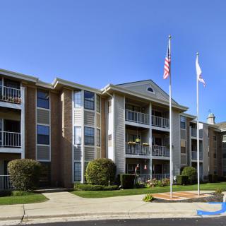 A three-story apartment building with balconies and flags flying in front, surrounded by well-maintained landscaping under a clear blue sky.