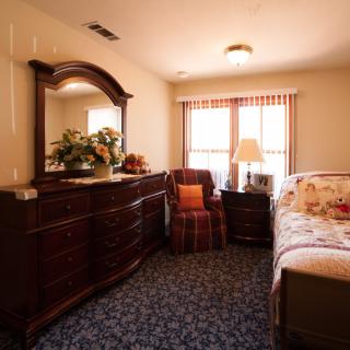 A cozy bedroom with wooden furniture, featuring a bed, dresser with mirror, and a lamp near the window.
