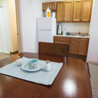 A cozy apartment kitchen featuring a wooden dining table set with a placemat and dishes, adjacent to cabinetry and a refrigerator.