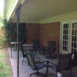A cozy patio area with outdoor furniture set beneath a covered space, featuring a brick wall and surrounded by greenery.