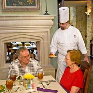 A chef presents a dish to a couple dining in a restaurant.