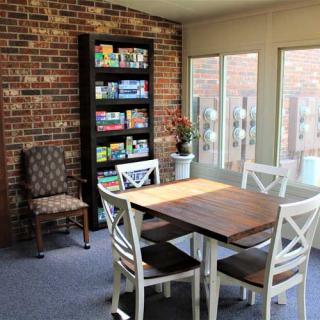 A cozy dining area with a wooden table, white chairs, and a bookshelf against a brick wall.