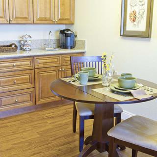 A cozy kitchen dining area featuring a wooden table set for two, surrounded by warm wooden cabinets.