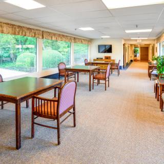 A bright dining area with tables and chairs, featuring large windows overlooking greenery.