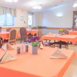 A bright dining room with tables set for a meal, featuring orange tablecloths and floral centerpieces.