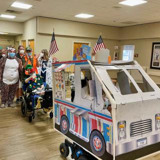 Residents enjoy a festive parade in a nursing home, showcasing colorful costumes and a creatively decorated vehicle.