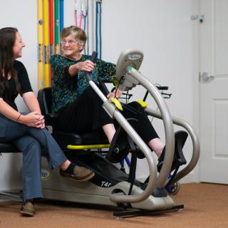 An elderly woman engages in a therapy session using a recumbent exercise bike, accompanied by a healthcare professional.