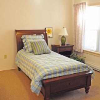 A cozy bedroom with a single bed and checkered bedspread, featuring a wooden nightstand with a lamp next to a window adorned with curtains.