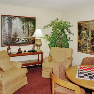 A cozy living room with comfortable chairs, a round table, decorative paintings, and a potted plant.