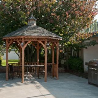 A cozy patio with a wooden gazebo and barbecue grill, surrounded by trees.