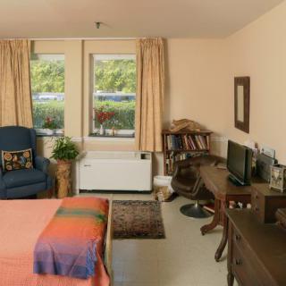 A cozy bedroom with a bed, armchair, and a desk, featuring large windows and tasteful decor.