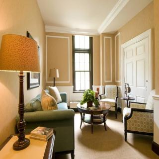 A cozy living room with elegant furnishings including a sofa, chairs, and a coffee table, warmly lit by a lamp next to a large window.