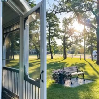 A serene view of a sunlit park from a porch, featuring a bench and surrounded by trees.