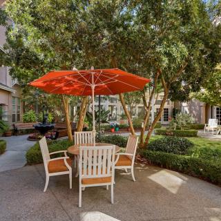 A tranquil patio setting with a table, chairs, and an orange umbrella surrounded by lush greenery.