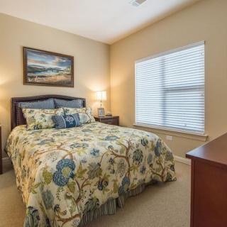 A cozy bedroom with floral bedding, featuring a window with blinds and a bedside lamp.