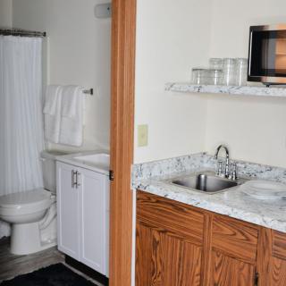 A small bathroom with an adjacent kitchenette featuring a sink, microwave, and shelves.
