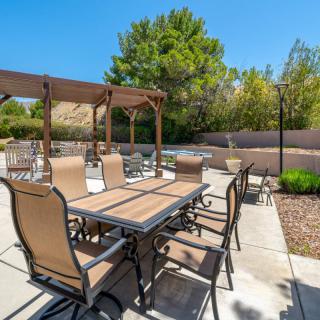 A sunny outdoor patio featuring a dining table with chairs, shaded by a pergola, surrounded by landscaped greenery.