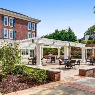 A well-maintained outdoor patio area featuring a white pergola, surrounded by lush greenery and adjacent to a brick building.