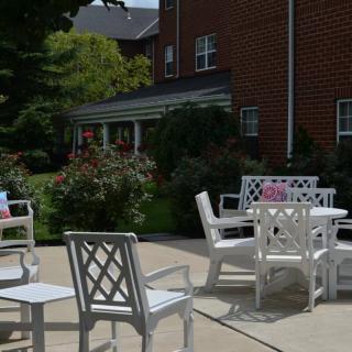A patio area with white furniture including chairs and a table, set against a brick building and surrounded by greenery.