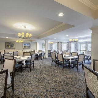 A spacious and elegant dining room with neatly arranged tables and chairs, well-lit by chandeliers and natural light from large windows.