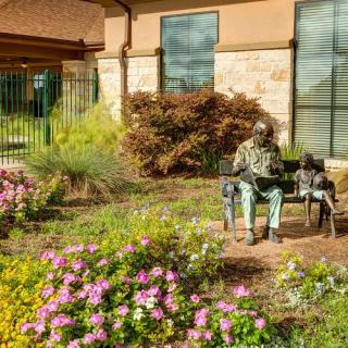 An elderly man sits on a bench surrounded by blooming flowers in a garden.
