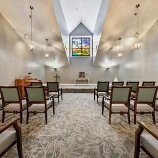 A serene chapel interior featuring rows of chairs, a stained glass window, and an altar at the front.