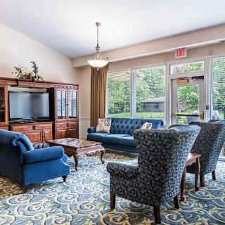 A cozy living room with blue sofas, patterned chairs, a wooden cabinet, and a television, illuminated by natural light from large windows.