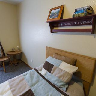 A cozy bedroom with a single bed, simple decor, and a wooden shelf displaying books and framed pictures.