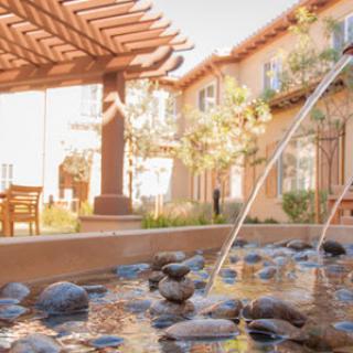 A serene outdoor space featuring a beautiful fountain surrounded by pebbles and shaded seating.