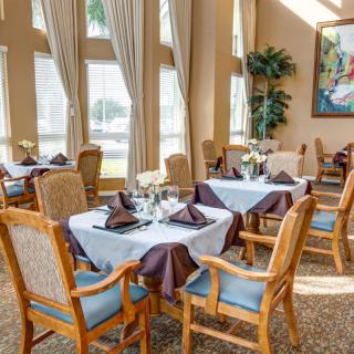 A well-lit dining room with elegantly set tables and wooden chairs, featuring large windows and tasteful decor.