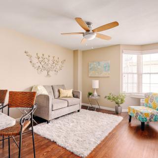 A cozy living room with a sofa, decorative wall art, and a ceiling fan, illuminated by natural light from large windows.