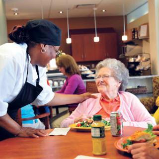 A friendly caregiver interacts with a senior diner, fostering a warm and welcoming atmosphere in a community dining setting.