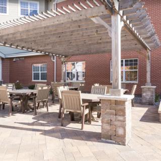 An outdoor patio with a pergola, featuring tables and chairs, adjacent to a brick building.