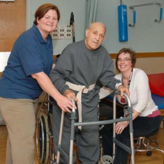 An elderly man receiving physical therapy support from two caregivers while using a walker.