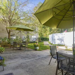 A sunny backyard patio with tables, chairs, and umbrellas surrounded by a garden.