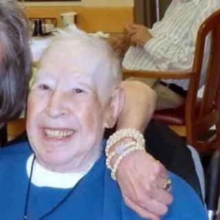 An elderly person smiling while seated indoors, wearing a blue shirt and pearl bracelet.