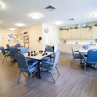 A well-lit community dining room with tables and chairs, featuring a small kitchen area.