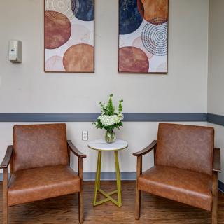 A cozy waiting area with two brown chairs, a small table adorned with flowers, and abstract artwork on the walls.