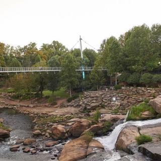 A scenic view of a bridge crossing over a river with waterfalls and surrounding greenery.