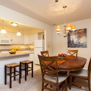 A modern dining area with a wooden table, chairs, and an open kitchen layout, featuring stylish lighting and bar stools.