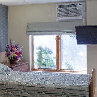 A cozy bedroom featuring a bed, vibrant flowers by the window, an air conditioning unit, and a mounted television.