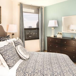 A cozy bedroom with elegant decor, featuring a bed with patterned bedding, a wooden dresser with lamps, and a window framed by curtains.