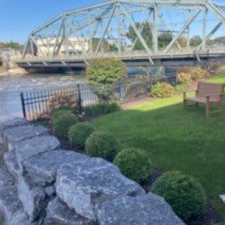 A scenic view of a bridge over a river, accompanied by a well-maintained garden with a stone wall and fencing on a sunny day.