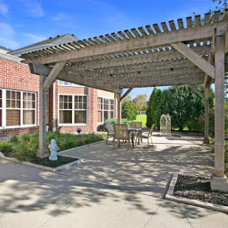 A cozy outdoor patio under a wooden pergola, surrounded by greenery and natural light.