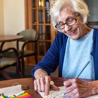 An elderly woman engaged in crafting at a table, smiling as she paints.
