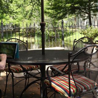 A cozy outdoor patio setting with a table and cushioned chairs under an umbrella, surrounded by greenery.