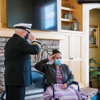 A military officer in uniform saluting a person seated in a wheelchair indoors, both wearing masks.